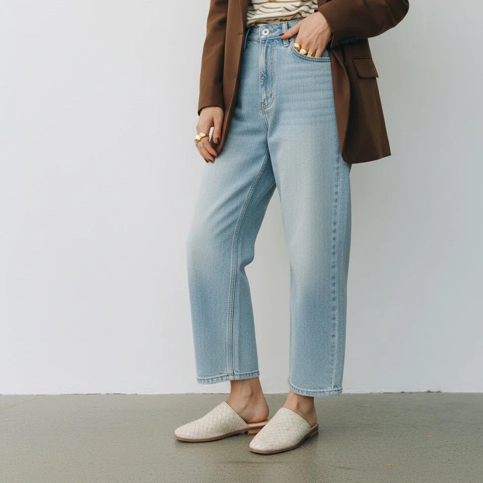A person wearing light-wash blue cropped jeans, a brown blazer, a striped top, and cream woven mule flats, standing against a plain light gray wall.