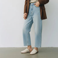 A person wearing light-wash blue cropped jeans, a brown blazer, a striped top, and cream woven mule flats, standing against a plain light gray wall.